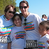 Four people wearing matching white The Color Run shirts and race bibs pose together outdoors, smiling at the camera. Two adults stand behind two children, celebrating inclusivity and raising awareness for hearing loss. Trees and other participants are visible in the background.