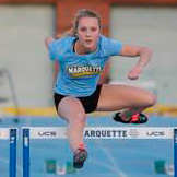 A female athlete with hearing loss in a blue Marquette shirt and black shorts leaps over a hurdle on an outdoor track during a race.