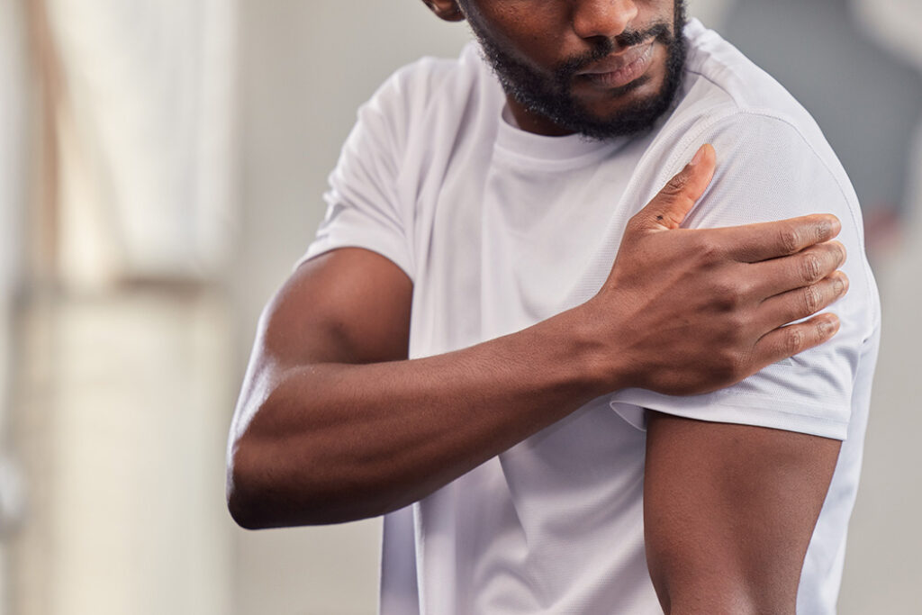 A man wearing a white t-shirt holds his left shoulder with his right hand, appearing to be in discomfort or pain—possibly experiencing symptoms related to hearing loss. The background is blurred.