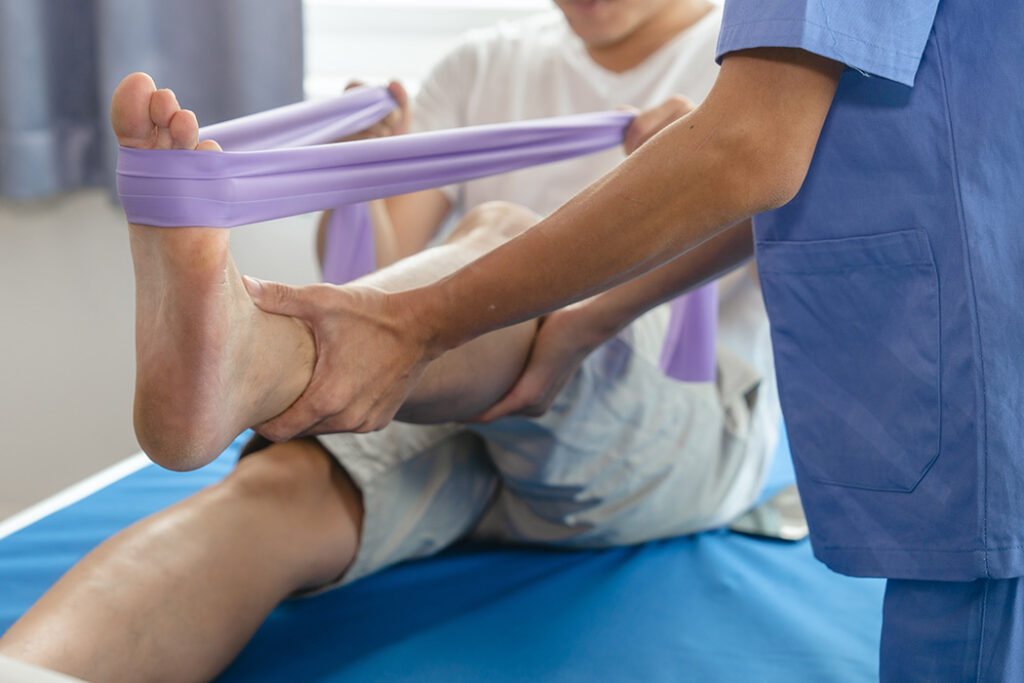 A physical therapist helps a patient exercise their leg using a purple resistance band while the patient, who has hearing loss, lies on a blue treatment table. The therapist supports the patient’s foot and ankle throughout the session.