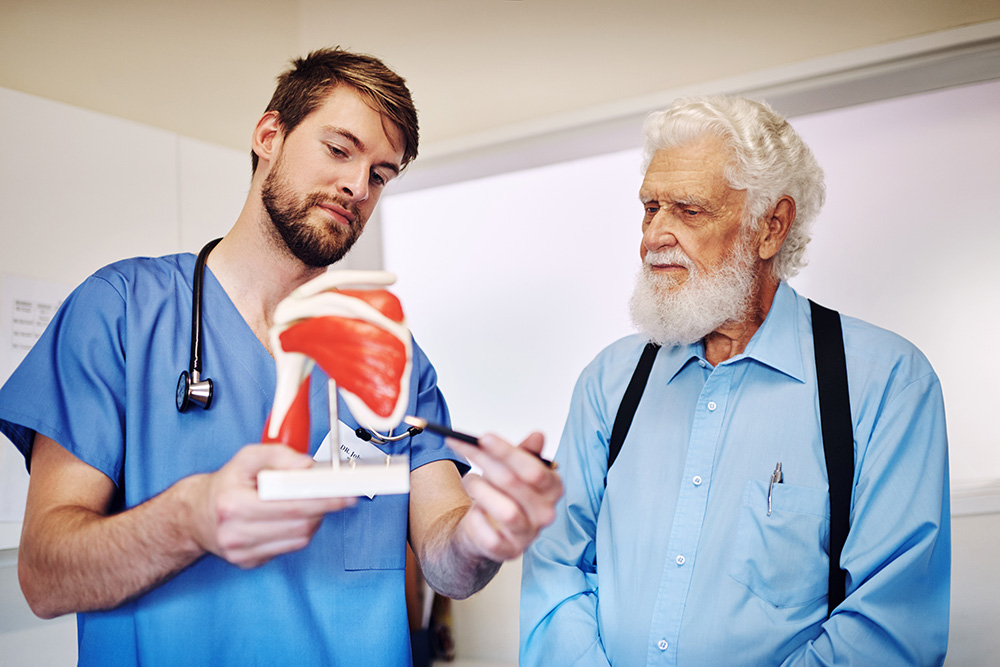 A healthcare professional in blue scrubs explains a shoulder joint model to an older man with white hair and a beard, both standing indoors.