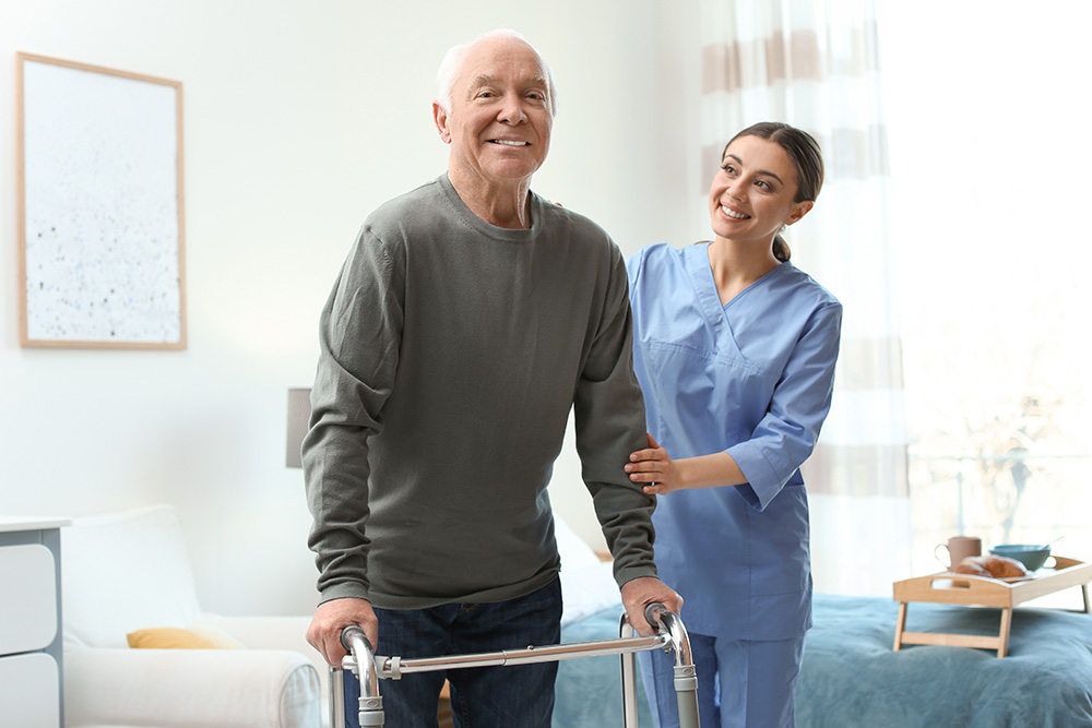 An elderly man using a walker smiles while a female caregiver in scrubs stands beside him, supporting him in a bright, tidy room with a bed and window in the background.