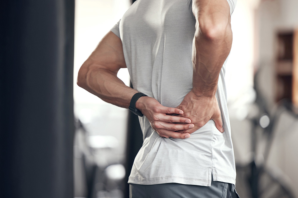 A person in a light gray shirt holds their lower back with both hands, appearing to be in pain, in what looks like a gym or fitness setting.