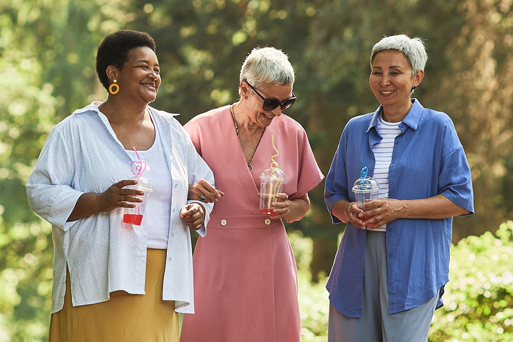 Three older women stand outdoors, smiling and laughing together. Each holds a clear cup with a colorful straw. They wear casual, brightly colored clothes and seem to be enjoying a sunny day in a green, leafy setting.