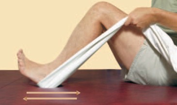 Person sitting on a red surface, using a white towel looped around their foot to gently pull their leg back with bent knee, suggesting a stretching or rehabilitation exercise. Two arrows indicate movement direction.