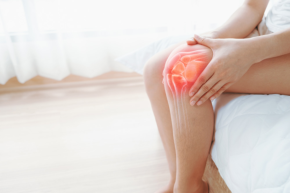 A person sitting on a bed holds their knee, which is highlighted with a red overlay and a transparent illustration of the knee joint, indicating pain or joint discomfort.