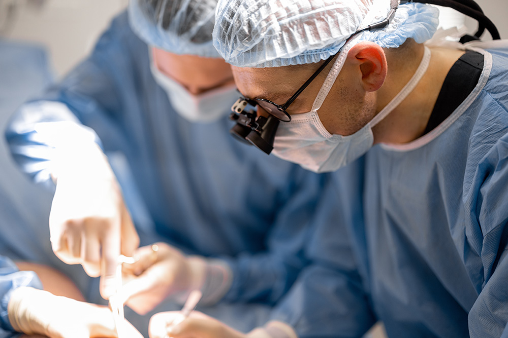 Two surgeons in blue scrubs, masks, and hair covers perform a procedure. One wears magnifying surgical loupes. Their hands are focused on the operation, with bright surgical lighting illuminating the area.