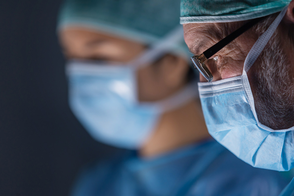 Two surgeons wearing surgical masks, caps, and scrubs are focused on their work. The foreground shows a close-up of one surgeon’s face, while the other appears blurred in the background.