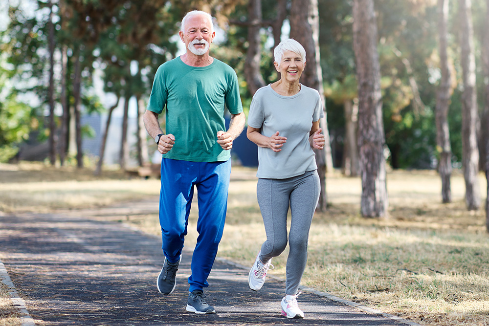 Two older adults, a man and a woman, are jogging together on a paved path in a park. They are smiling and dressed in comfortable athletic wear, surrounded by trees and nature.