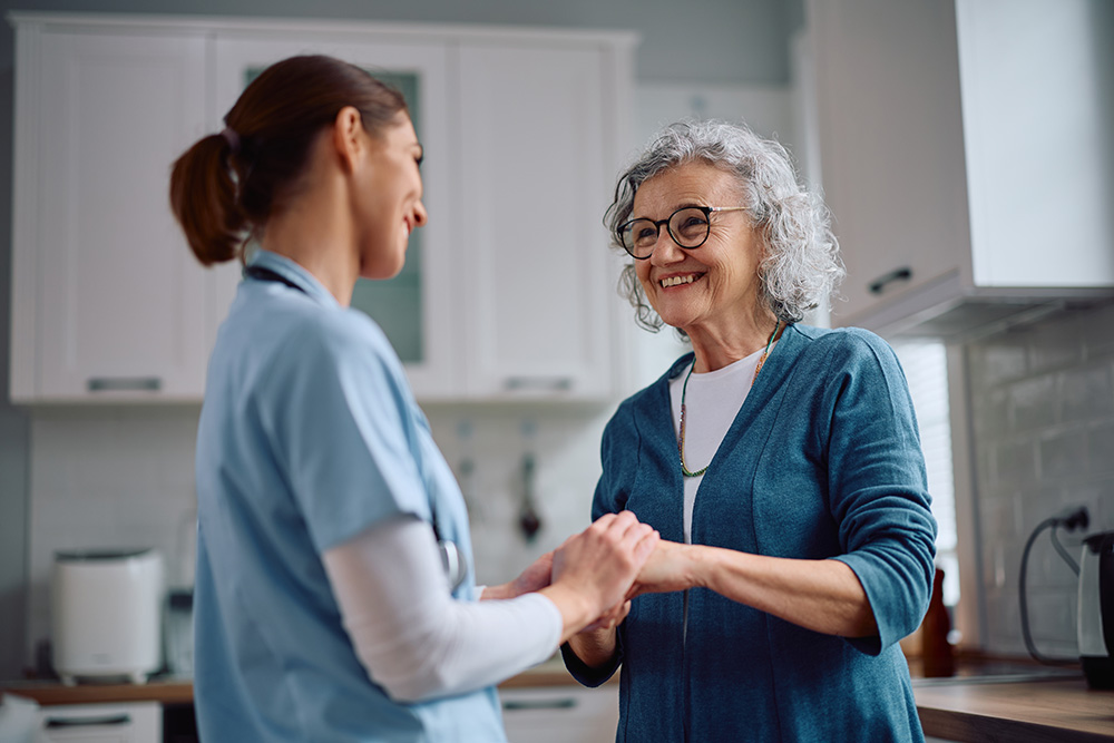 A smiling older woman with gray hair and glasses holds hands with a nurse in blue scrubs in a bright kitchen, conveying warmth and support.