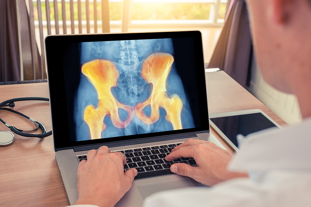 A person uses a laptop displaying a colored X-ray image of the pelvic bones. A stethoscope and a tablet are on the wooden desk nearby.