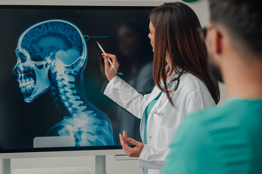 A doctor in a white coat points at a digital X-ray image of a human skull on a screen, explaining details to another person in medical scrubs.