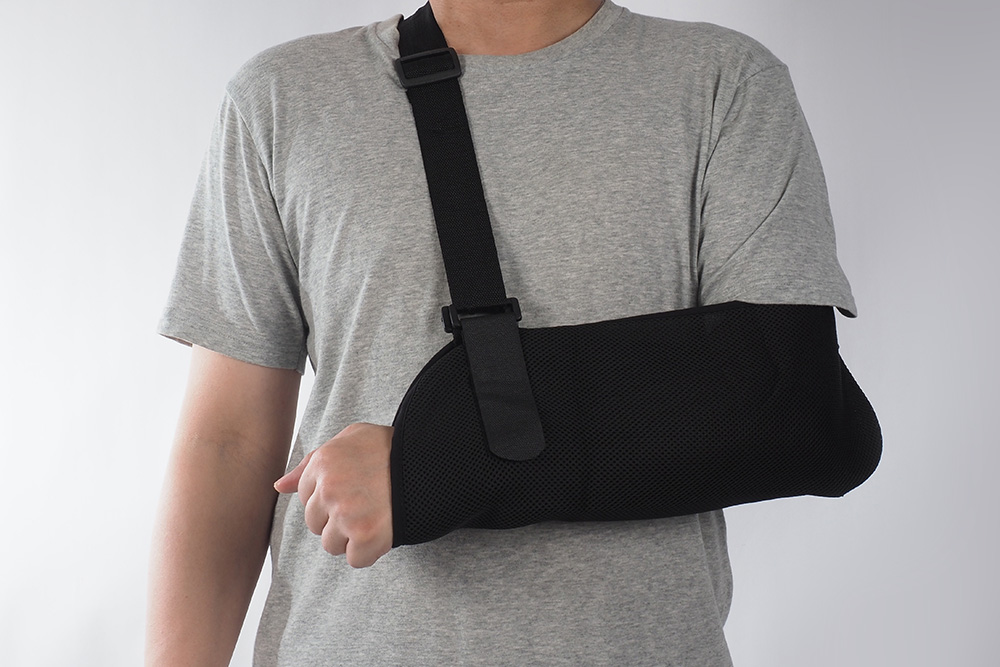 A person wearing a grey T-shirt has their right arm in a black fabric arm sling, secured with a strap around their neck, against a plain light background.