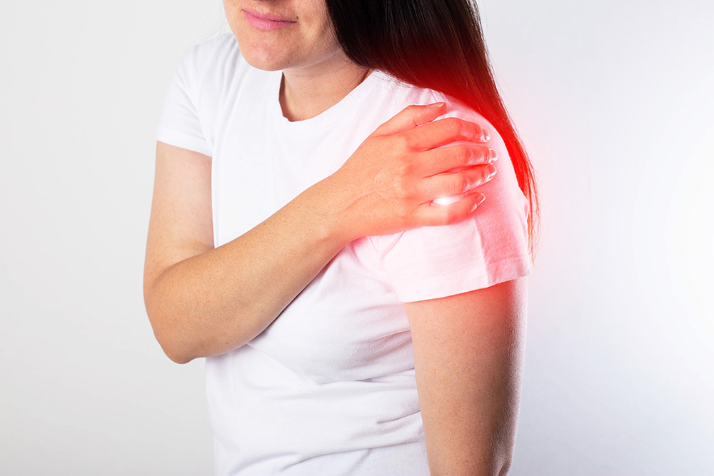 A woman in a white t-shirt touches her left shoulder, which is highlighted with a red glow, indicating pain or discomfort in that area.