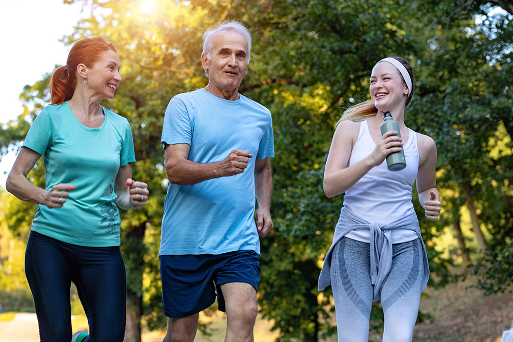 Three people, two women and one man, are jogging together outdoors in a park on a sunny day. They appear happy and are smiling, with trees and greenery in the background. One woman holds a water bottle.
