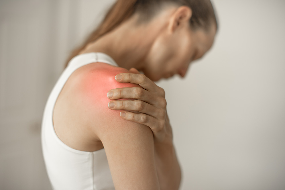 A woman in a white tank top holds her left shoulder, which appears red and inflamed, indicating pain or injury. She is looking down with a pained expression.