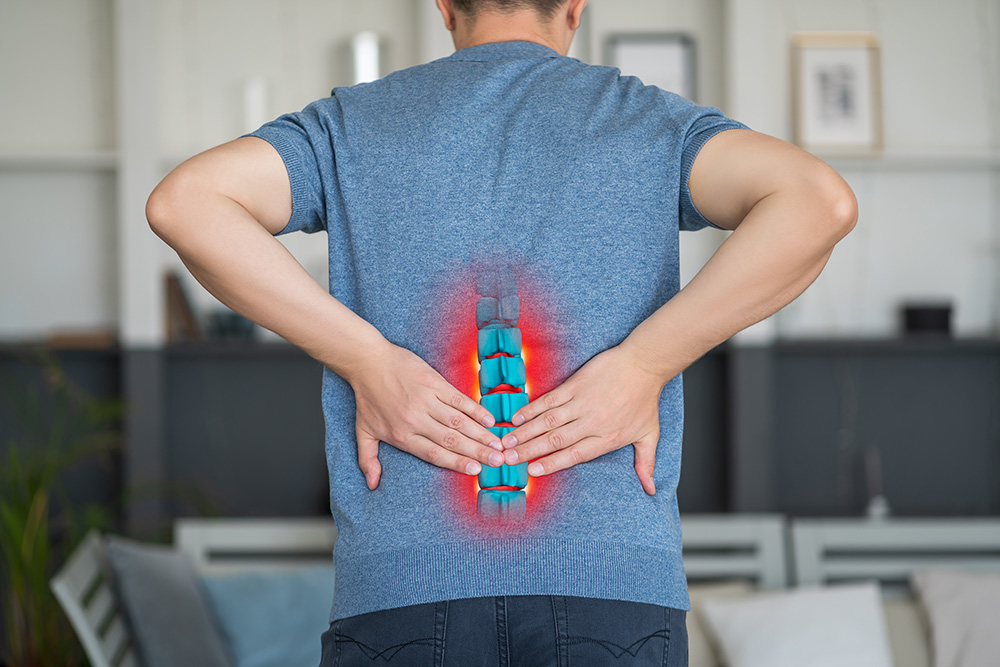 A person in a blue shirt holds their lower back with both hands. An illustration of a highlighted spine with red pain marks is superimposed on the lower back area, indicating discomfort or back pain.
