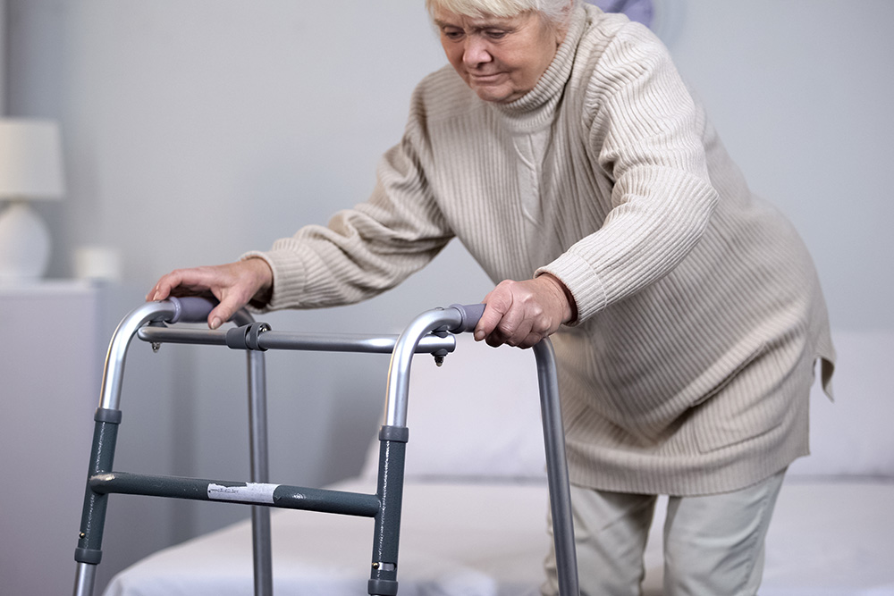 An elderly woman wearing a beige sweater uses a walker for support as she stands up from a bed in a softly lit room.