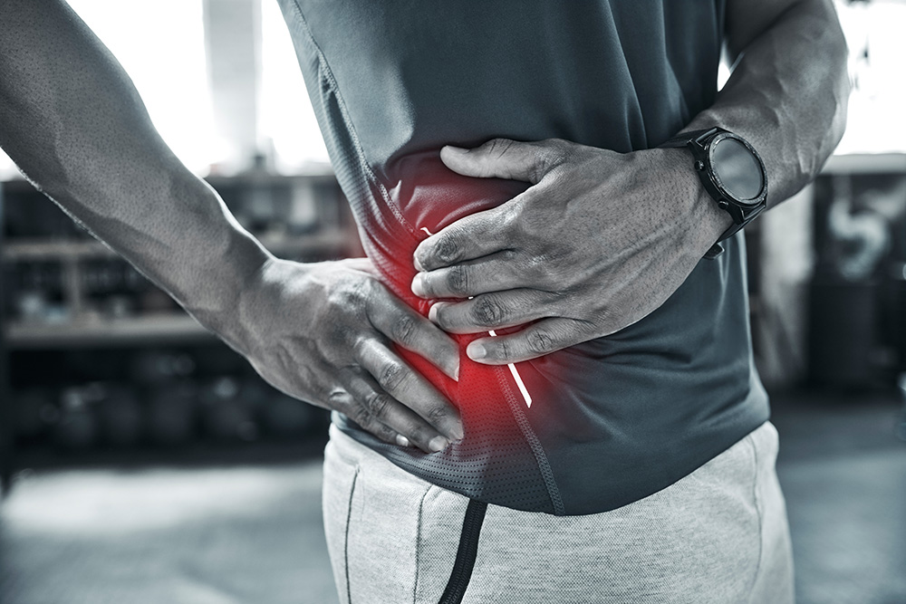 A person wearing athletic clothes holds their lower back in pain, with a red highlight indicating the painful area. The setting appears to be a gym or fitness center.