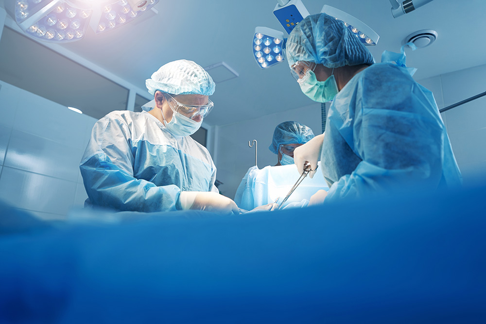 Surgeons in blue scrubs, masks, and protective eyewear perform an operation under bright surgical lights in a sterile operating room. Medical instruments and equipment are visible.