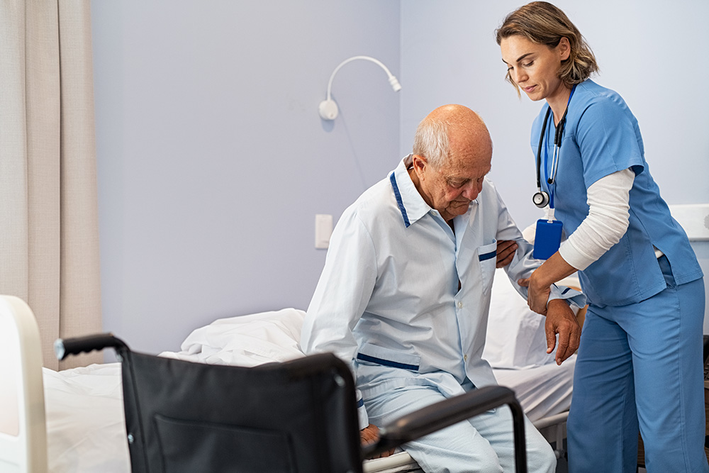 A nurse in blue scrubs assists an elderly man in pajamas as he sits on the edge of a bed, preparing to move him toward a nearby wheelchair in a hospital or care facility room.