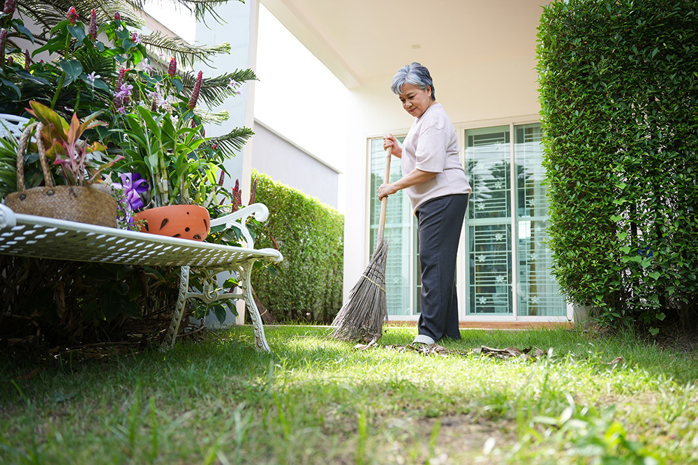 An older woman with short gray hair is raking leaves in a garden beside a white bench and potted plants, near a house with large glass doors and green hedges.