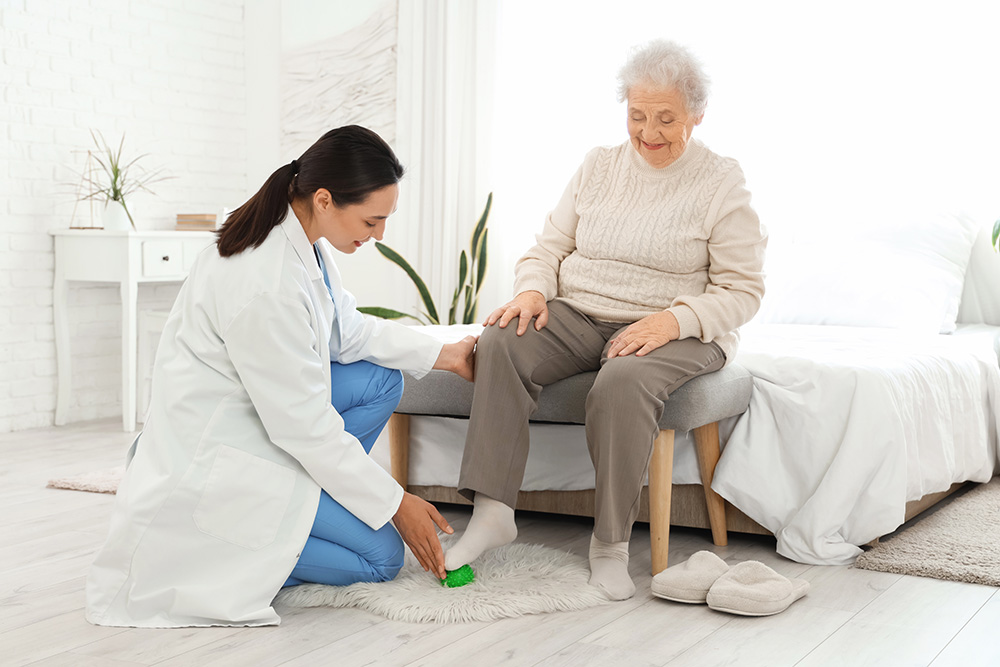 A female doctor examines the foot of an elderly woman who is sitting on a bed. The elderly woman is smiling, and the doctor is kneeling on the floor, holding the patient’s ankle. Slippers are placed on the floor nearby.