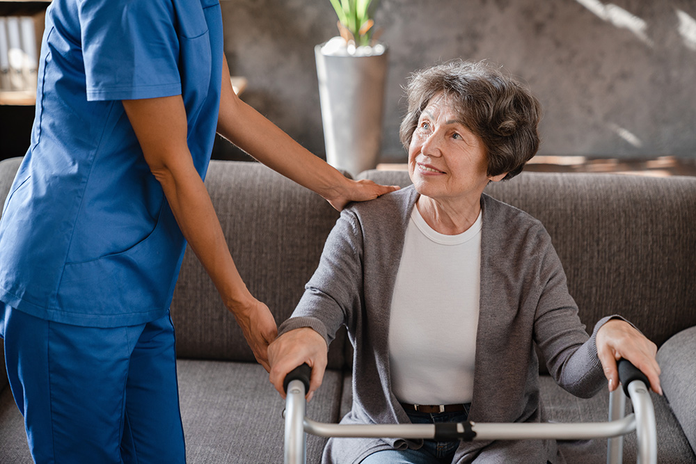 An older woman using a walker sits on a couch, looking up and smiling at a caregiver in blue scrubs, who is gently touching her shoulder and offering support.