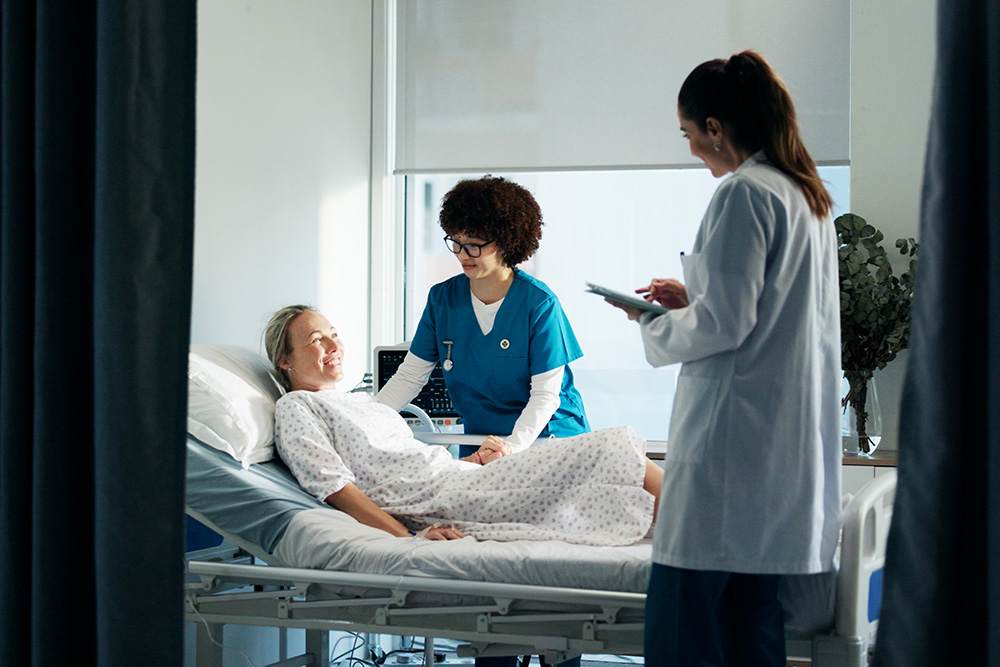 A patient lies in a hospital bed smiling at a nurse standing beside her, while a doctor stands nearby holding a clipboard and taking notes. The room is bright with natural light.