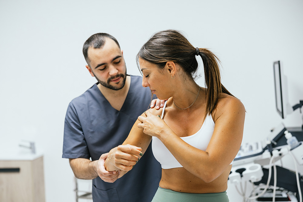 A healthcare professional in gray scrubs examines a womans shoulder and arm in a medical office. The woman, wearing a sports bra and leggings, appears to be describing pain or discomfort. Medical equipment is visible in the background.