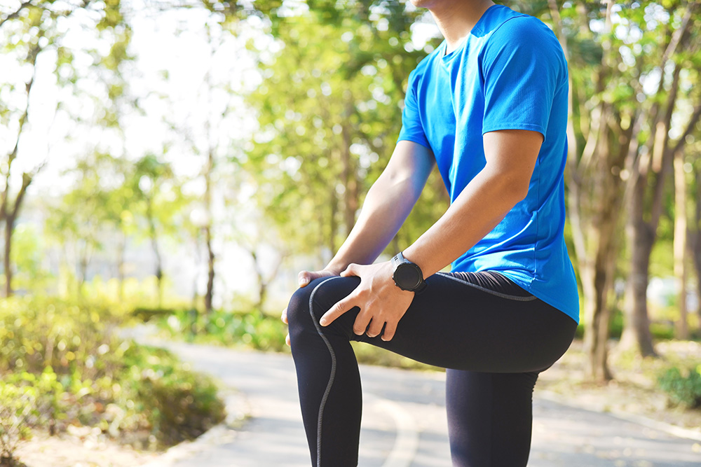 A person in a blue shirt and black leggings stretches their leg and holds their knee while standing on a winding path in a sunny, tree-filled park. Their face is not visible.