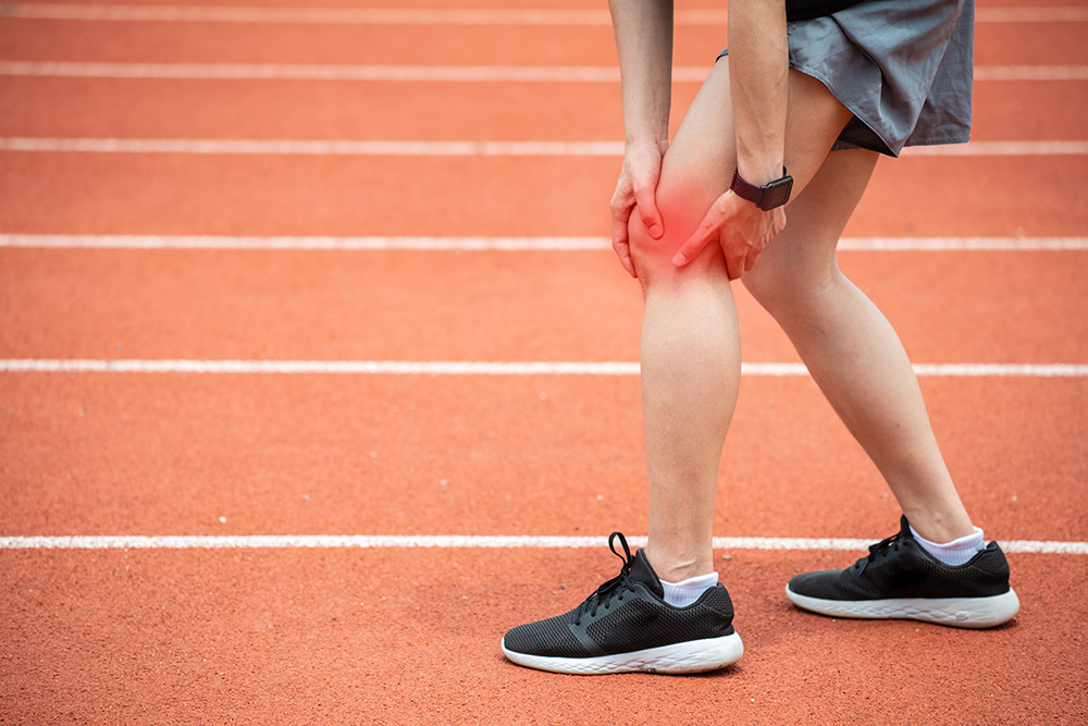 A person wearing athletic shoes and shorts is holding their knee in pain while standing on a running track, suggesting a possible sports injury.
