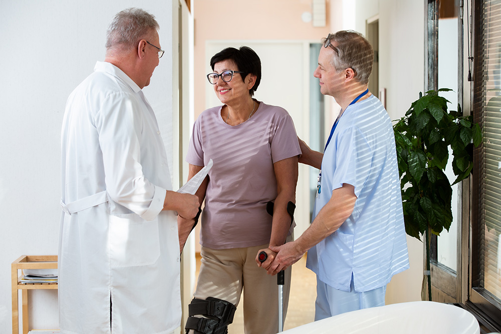 A woman with a leg brace and crutches smiles while talking to two healthcare professionals in a bright medical clinic. One professional holds her arm for support, and the other holds a clipboard.