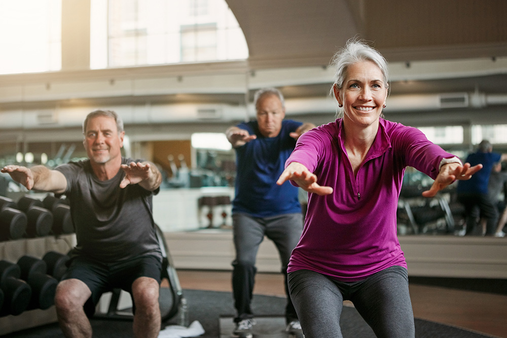 Three older adults exercise indoors, performing squats with arms extended forward. The group appears energetic and focused, with a woman in a purple top smiling at the front of the class. Mirrors and gym equipment are in the background.