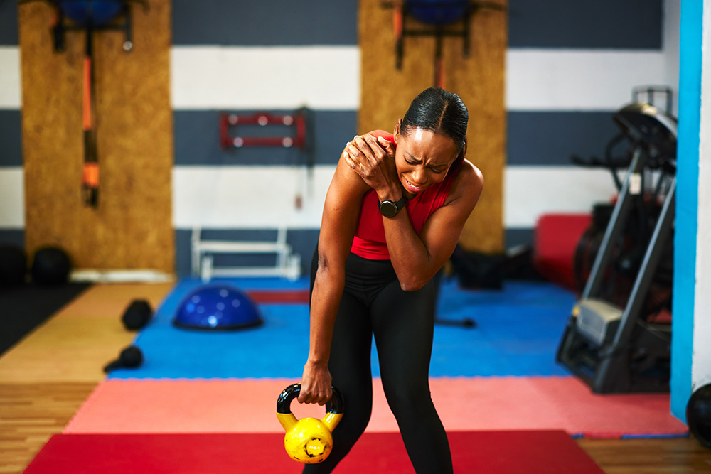 A woman in athletic wear holds her shoulder in pain while lifting a yellow kettlebell in a gym, appearing to have an injury during her workout.