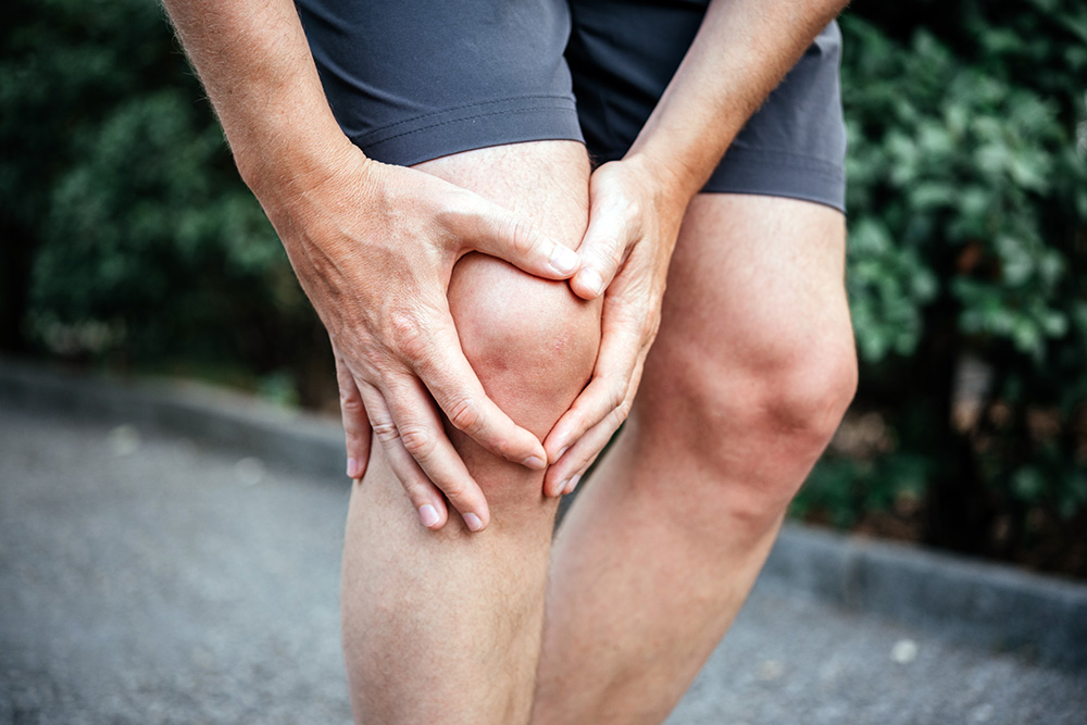 A person wearing gray shorts is holding their left knee with both hands, appearing to be in pain while standing outdoors on a path with greenery in the background.