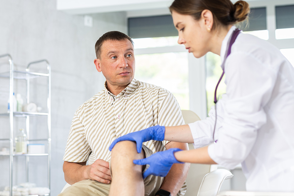 A healthcare professional in gloves examines a seated mans knee in a medical office. The man looks at the examiner with a concerned expression. Medical equipment is visible in the background.