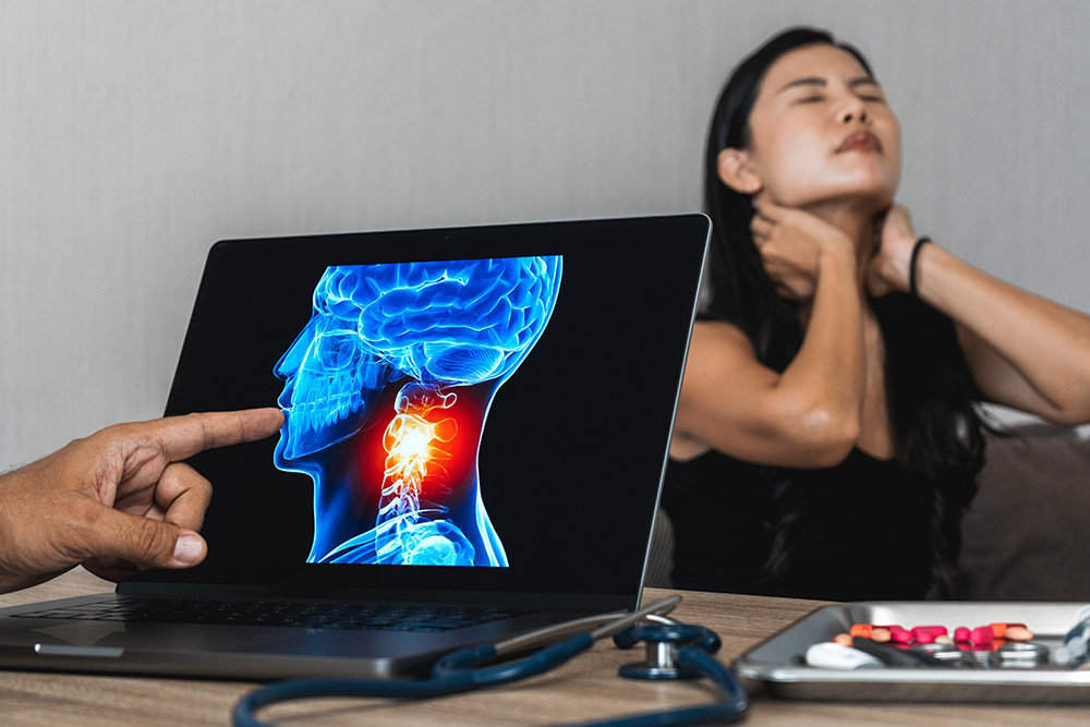 A person points to a neck injury on a medical illustration displayed on a laptop, while a woman in the background holds her neck in pain. A stethoscope and pills are on the table.