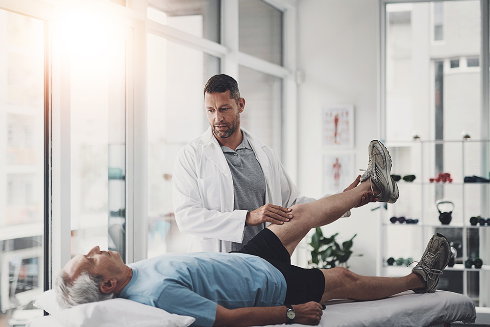 A male doctor or physical therapist in a white coat examines an older man’s leg as he lies on an exam table in a bright, modern clinic. The therapist lifts the patient’s leg, assessing movement and flexibility.