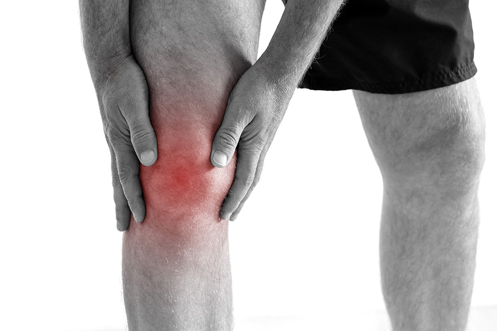 A person in black shorts holds their knee with both hands, highlighting pain or injury. The knee area is shown in red to indicate inflammation or discomfort.