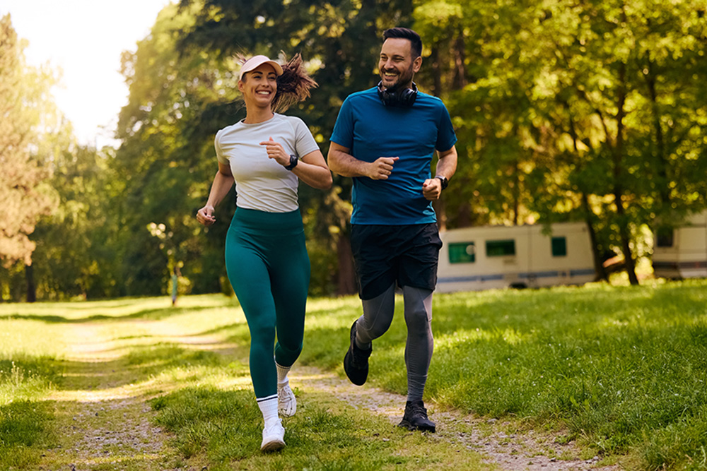 A man and woman jog together on a sunny path in a park surrounded by trees. Both are smiling and wearing athletic clothing, and the background shows a trailer among the greenery.
