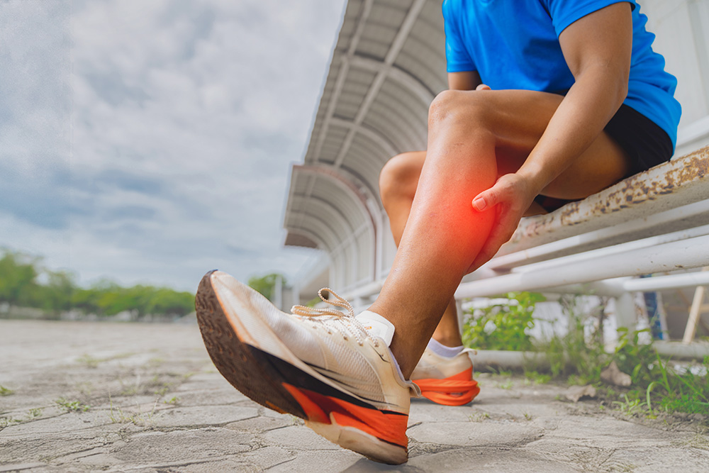 A person in sportswear sits on a bench outdoors, holding their lower leg in pain. The calf area is highlighted in red, indicating discomfort or injury.