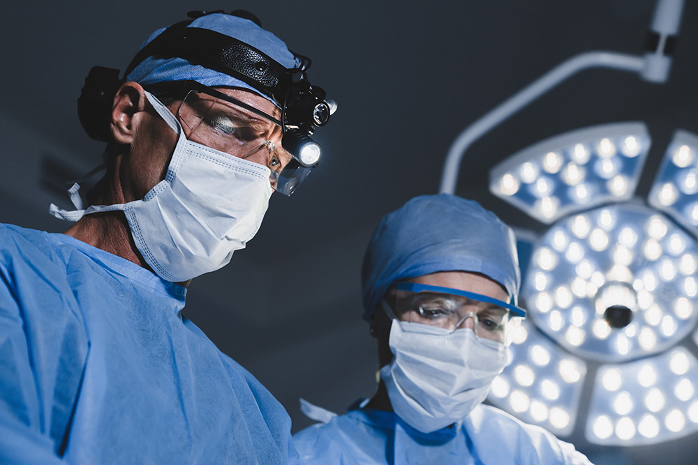 Two surgeons wearing blue surgical gowns, masks, and headlamps focus on a procedure under bright operating room lights. Medical equipment and a lamp are visible in the background.