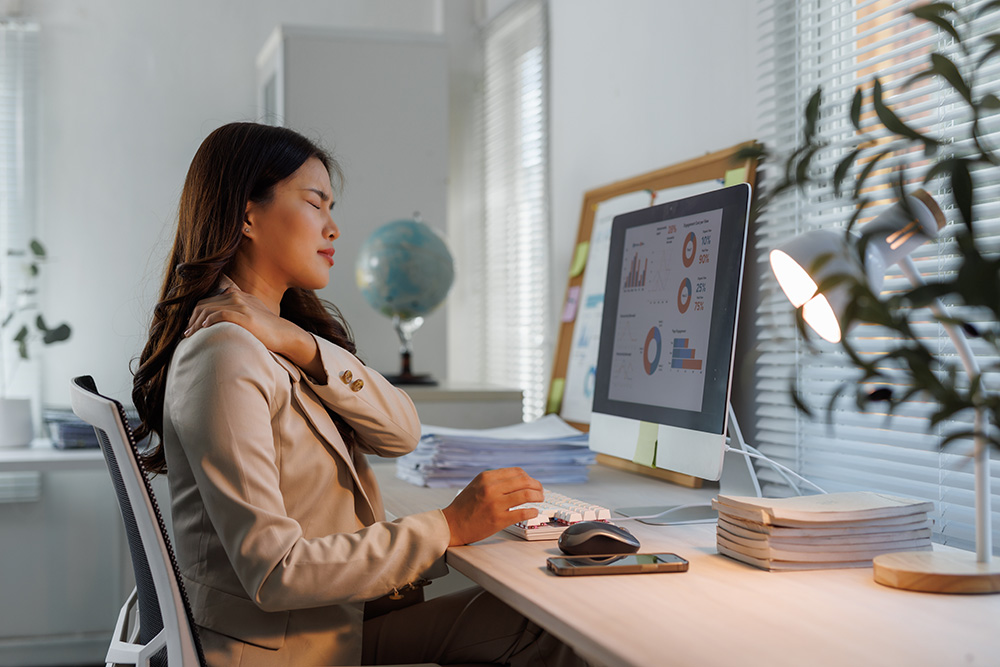 A woman in business attire sits at a desk, touching her shoulder with a pained expression. A computer screen displays charts and graphs. The office is well-lit with sunlight through blinds and has neatly arranged papers and plants.