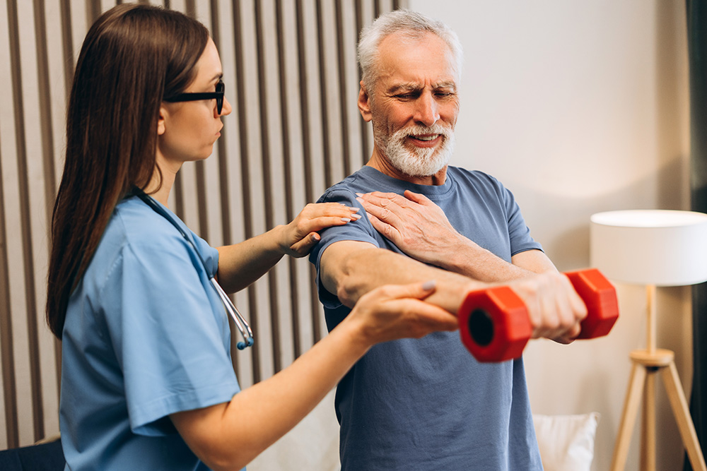 A nurse helps an older man in a blue shirt lift a red dumbbell while he holds his shoulder in discomfort, guiding him through a physical therapy exercise.