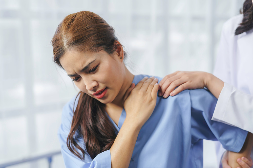 A woman in a blue hospital gown holds her shoulder in pain while a healthcare professional places a comforting hand on her other shoulder.