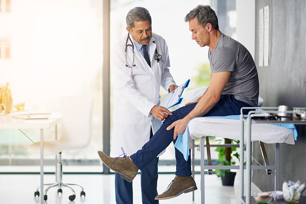 A doctor in a white coat examines a middle-aged mans knee as the man sits on an exam table in a bright medical office. The doctor holds a clipboard and appears to be discussing the patients condition.