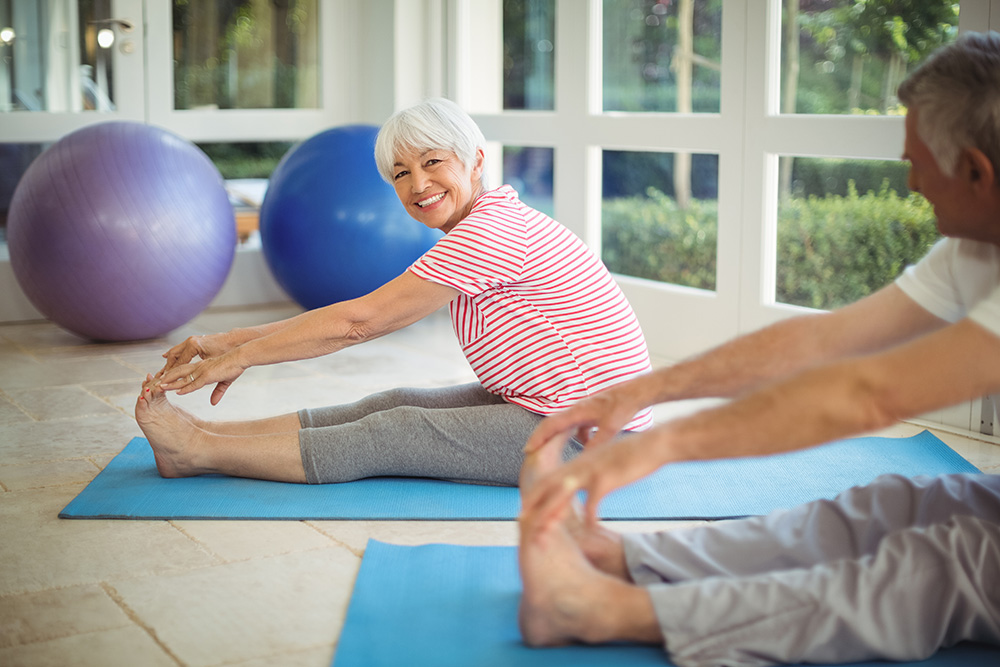 An older woman and man sit on yoga mats indoors, stretching forward to touch their toes. The woman is smiling at the camera. Large exercise balls and windows with a garden view are in the background.