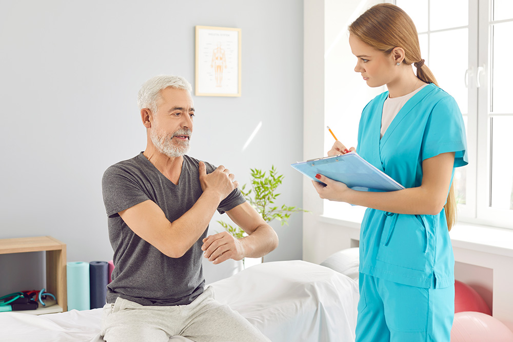An older man sitting on an exam table points to his shoulder while a female healthcare professional in blue scrubs takes notes on a clipboard in a bright medical office.