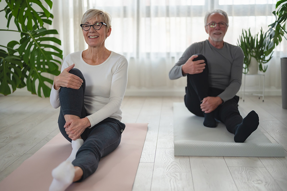 Two older adults sit on yoga mats indoors, smiling as they stretch one knee to their chest. Large green plants and sheer curtains are visible in the bright, airy room.